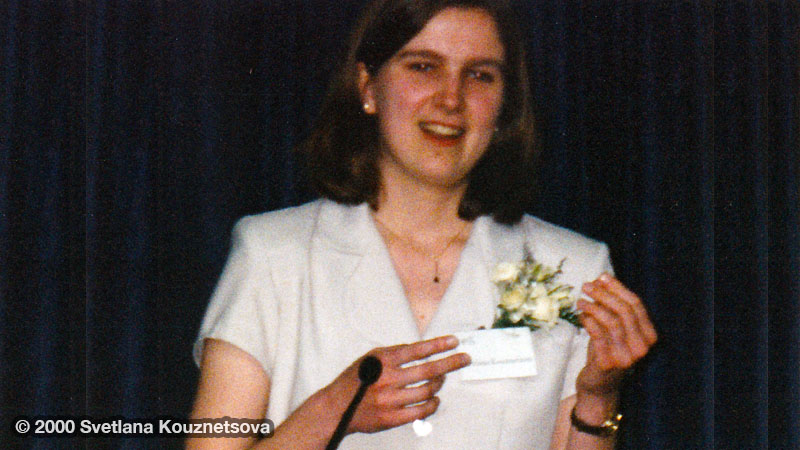 A brunette in a short sleeved beige jacket with a small flower bouquet pinned to it giving a speech.