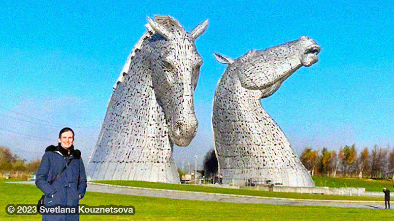 A brunette in a brown coat standing in front of 2 large metallic horse heads.